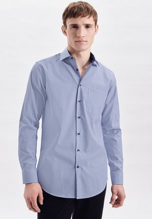 Jeune homme portant une chemise habillée bleu clair à manches longues, boutonnée, avec de fines rayures verticales et une poche poitrine, debout devant un fond uni.