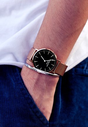 Cadran noir avec de fins repères blancs, boîtier rond en or et bracelet en maille rose, porté sur un poignet, accompagné d'un bracelet en métal.