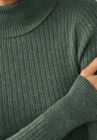 Pull à col roulé côtelé en tissu maille vert foncé, avec des manches longues et un motif texturé. Vue en gros plan montrant le détail de la manche.
