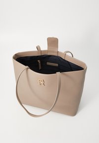 Sac fourre-tout beige en cuir synthétique, avec un logo en métal doré, deux longues anses et un intérieur sombre avec une poche zippée.