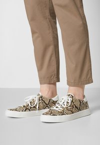 Zapatillas con un estampado de piel de serpiente en tonos marrones y beige, que cuentan con cordones blancos y una suela de goma blanca con acentos de hardware dorado.