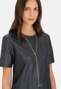 Guldkæde med forlængede led, udstyret med en toggle-lås, båret over en mørk lædertop med en struktureret design.