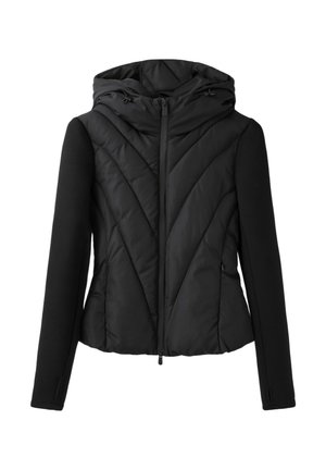 Femme debout sur fond gris portant une veste à capuche noire, un haut à manches longues noir, des leggings noirs et des bottes noires montantes.