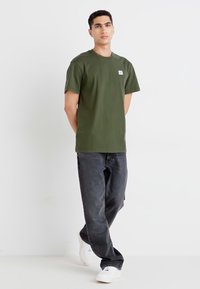 Olijfgroene t-shirt met korte mouwen, een rechte pasvorm en een klein merkteken op de borst, gecombineerd met donkere jeans en witte sneakers.