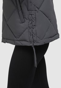 Gray quilted puffer jacket featuring lace-up detail on the side and a drawstring at the hem, paired with black textured leggings.