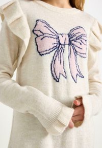 Lys cremesviter med flæseeffekter på ærmerne, prydet med et strikket bue-design i pink og marineblå på forsiden. Blød tekstur, ribbede manchetter.