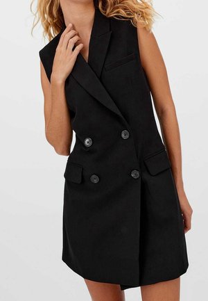 Robe blazer noire sans manches, croisée, avec des revers et des poches à rabat, portée par une personne aux cheveux bouclés blonds.