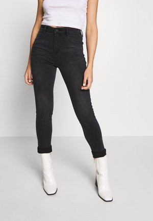 Personne portant un jean skinny noir taille haute retroussé aux chevilles et des bottines blanches à talons, associé à un haut blanc sans manches.