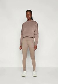 Sportos öltözék, amely egy rövidített barna pulóvert és az ehhez illő, oldalsó zsebekkel ellátott testhez álló leggingset tartalmaz, fehér sportcipővel párosítva.