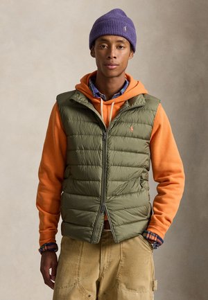 Jonge man die een paarse gebreide muts, oranje hoodie, groene doorgestikte vest, geruit overhemd, beige broek en een polshorloge draagt tegen een neutrale achtergrond.