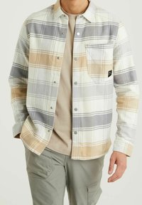 Homme portant une chemise à carreaux beige et grise boutonnée par-dessus un t-shirt beige avec un pantalon cargo vert, debout, les mains relaxées le long du corps.