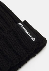 Black knitted beanie with a textured ribbed design. Features a rectangular label with "manastash" in white text on a black background.