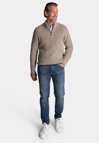Beige gebreide pullover met een ritssluiting bij de nek, gecombineerd met blauwe denim jeans en witte sneakers. Het ontwerp heeft een geribbelde textuur en een klassieke pasvorm.