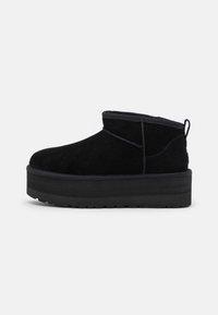 Botas de tobillo de ante negro con punta redonda, adornos cosidos y suela de plataforma negra gruesa. Presenta una lengüeta en la parte posterior.