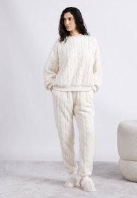 Γυναίκα που στέκεται με τα χέρια στις τσέπες, φορώντας υφασμάτινη λευκή άνετη ενδυμασία και παντόφλες, σε γκρι μοκέτα δίπλα σε μαξιλάρι σκαμπό.