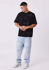 Zwart T-shirt met witte sterontwerpen, gecombineerd met lichtblauwe, versleten jeans met soortgelijke sterpatronen, aangevuld met grijze sneakers.
