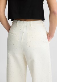 Široké krémové kalhoty z denimu s rozházenými kamínkovými ozdobami a měkkým elastickým pasem. Obsahují dvojité zadní kapsy.