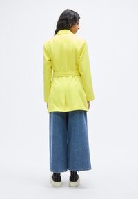 Κίτρινο σακάκι με ζώνη, κατασκευασμένο από λείο ύφασμα, που διαθέτει γιακά και πίσω άνοιγμα. Συνδυάζεται με μπλε τζιν παντελόνι φαρδιάς γραμμής και μαύρα παπούτσια.