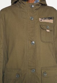 Chaqueta con capucha de color verde oliva hecha de un tejido texturizado; presenta botones grandes, bolsillos frontales y una etiqueta con costura en contraste.
