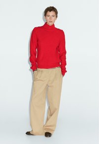 Červený žebrovaný rolák s knoflíky na rameni, spárovaný s béžovými širokými kalhotami a tmavými botami, pózující na světlém pozadí.