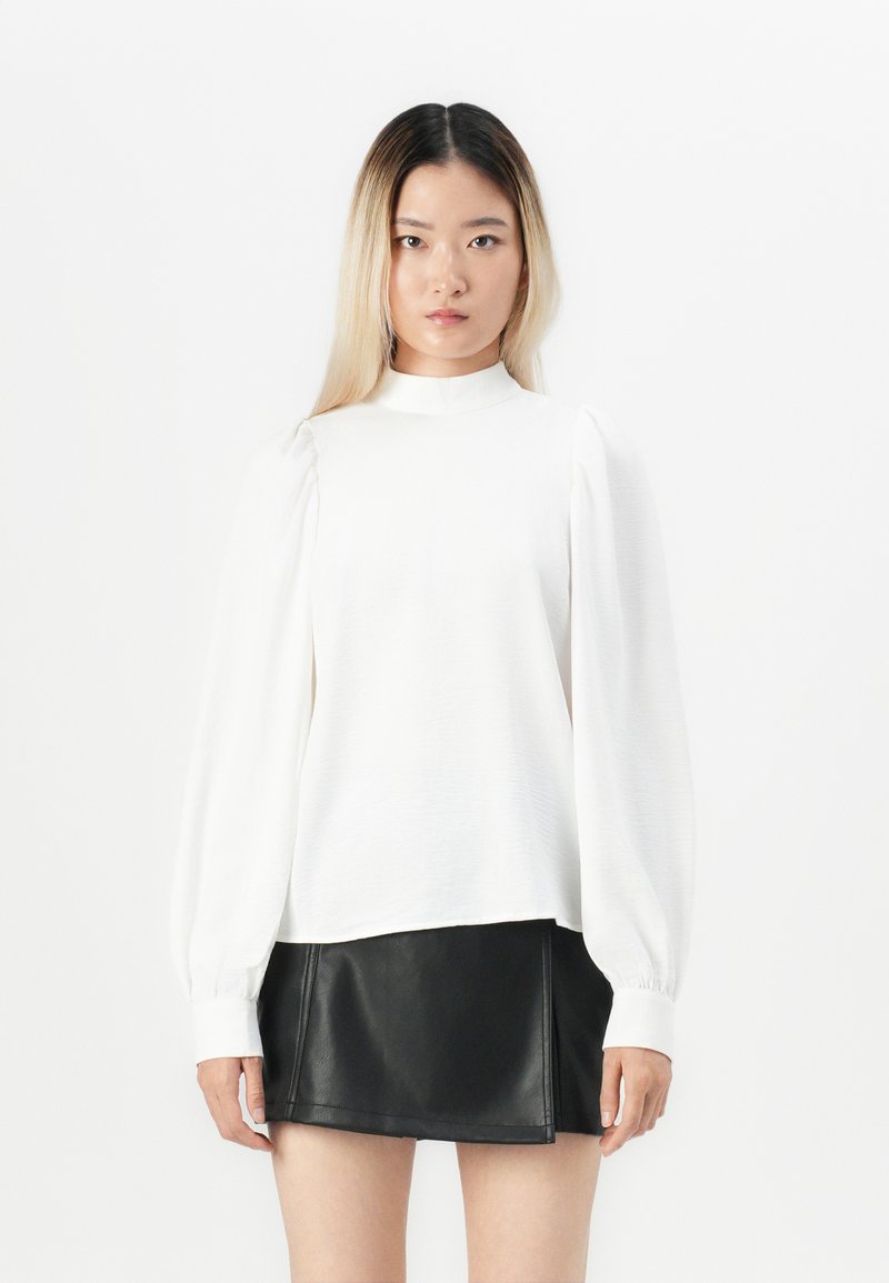 White long-sleeve top with a high neckline and textured fabric, paired with a fitted black leather mini skirt.