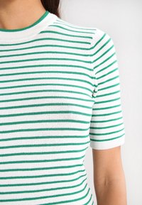 Pruhovaný pletený top se zelenými a bílými vodorovnými pruhy, krátkými rukávy a kulatým výstřihem se zeleným kontrastním lemem.
