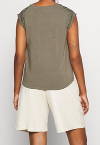 Person iført en olivengrønn ærmeløs top med blonde skuldre og cremede knælange shorts, set fra ryggen, der viser en sol tatovering på armen.