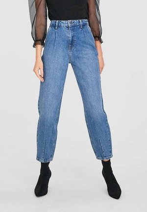 Kobieta nosząca niebieskie dżinsy z wysokim stanem z plisami z przodu, czarną przezroczystą bluzkę z długim rękawem oraz czarne botki na obcasie.