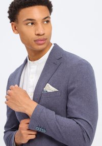 Marineblauwe blazer met een gestructureerde afwerking, met daaronder een wit overhemd en een gestreepte pochette in tinten van crème en blauw.