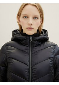 Giubbotto a vento nero con motivo a chevron, chiusura con zip frontale e cappuccio. Il materiale ha una finitura lucida e presenta un colletto alto per il calore.