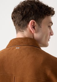 Brown suede jacket with a folded collar and visible stitching. Features a small "BOSS" logo near the neckline and smooth texture.