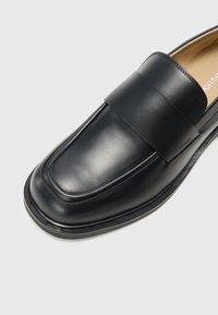 Crne kožne loaferice s glatkom površinom, širokom trakom preko prednjeg dijela, zaobljenim nosom i izdržljivim potplatom. Minimalno brendiranje sa strane.