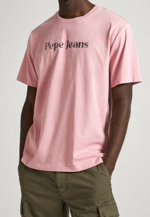 Homme portant un t-shirt rose à manches courtes avec le texte « Pepe Jeans » et un pantalon cargo vert olive, les mains dans les poches, sur un fond uni.