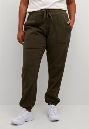 Personne portant un pantalon de jogging vert olive avec cordon et poches avant, et des baskets blanches, debout devant un fond uni.
