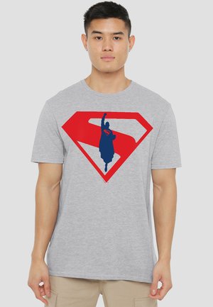 Young man wearing grey t-shirt with a red Superman emblem featuring a blue silhouette of a flying figure with raised fist.