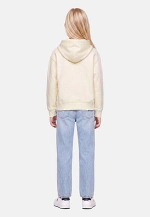 Barn med langt blondt hår iført en cremefarvet hættetrøje, lyseblå jeans og hvide sneakers står med ryggen mod en ensfarvet baggrund.