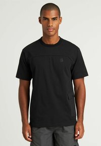 T-shirt nera a maniche corte con scollatura a girocollo, che presenta un pannello testurizzato sul petto e una tasca laterale con chiusura a bottone.