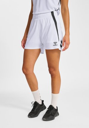 Person iført hvide atletikshorts med sorte striber, hvide sokker og sorte sneakers, stående mod en ensfarvet baggrund.