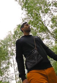 Felpa con cappuccio nera, pantaloni arancioni e una borsa a tracolla in rete nera. Circondato da alberi verdi e un cielo nuvoloso.