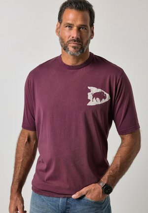 Mand med skæg iført en vinrød t-shirt med hvidt bjørnemotiv på brystet, blå jeans og sort ur, stående foran en ensfarvet baggrund.