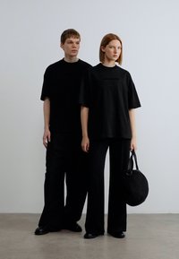 Δύο άτομα φορούν ίδια φαρδιά μαύρα μπλουζάκια και άνετα μαύρα παντελόνια. Το ένα κρατάει μια στρογγυλή μαύρη τσάντα από λείο ύφασμα με ελάχιστες λεπτομέρειες στο σχέδιο.