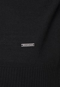 Tissu tricoté noir avec une petite étiquette rectangulaire en métal portant la mention « JOOP! » cousue près du centre.