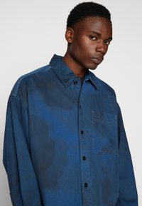 Blå skjorte med krage som kan knappes ned, med en ensfarget mønster og en brystlomme. Laget av teksturert stoff med en myk overflate.