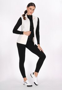 Vesta albă din fleece fără mâneci, cu guler înalt și fermoar în față; asortată cu un top negru cu mâneci lungi și colanți negri; adidași albi.