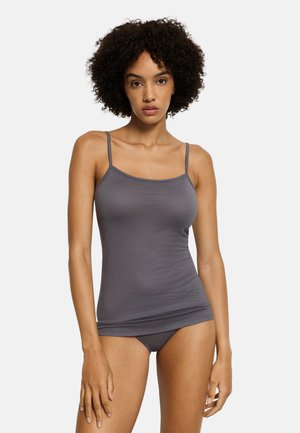 Frau mit lockigem Haar, die ein eng anliegendes graues Camisole und passende Unterwäsche trägt, steht vor einem schlichten hellen Hintergrund.