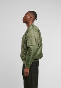 Giubbotto bomber verde oliva con finitura satinata, polsini a coste, tasca frontale e tasca laterale con zip, indossato con pantaloni neri.
