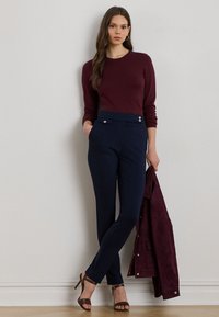 Žena v bordovom svetri, tmavomodrých nohaviciach a hnedých lodičkách, ktorá drží zodpovedajúcu bordovú bundu a stojí pri svetlej stene.