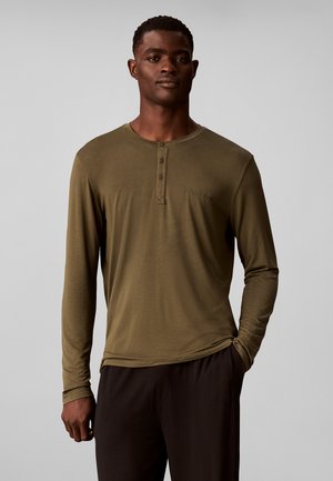 Homme portant un t-shirt henley manches longues vert olive et un pantalon foncé, debout devant un fond gris uni avec une main dans la poche.