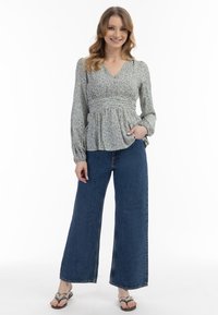Blumenmuster-Bluse mit langen Ärmeln und gerafftem Taillenbereich in hellgrün, kombiniert mit weit geschnittenen blauen Jeans. Modell trägt flache Sandalen.