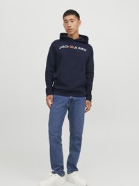 Sweat à capuche marine avec une poche avant et un logo blanc, associé à un jean en denim bleu et des baskets blanches, posé sur un fond neutre.
